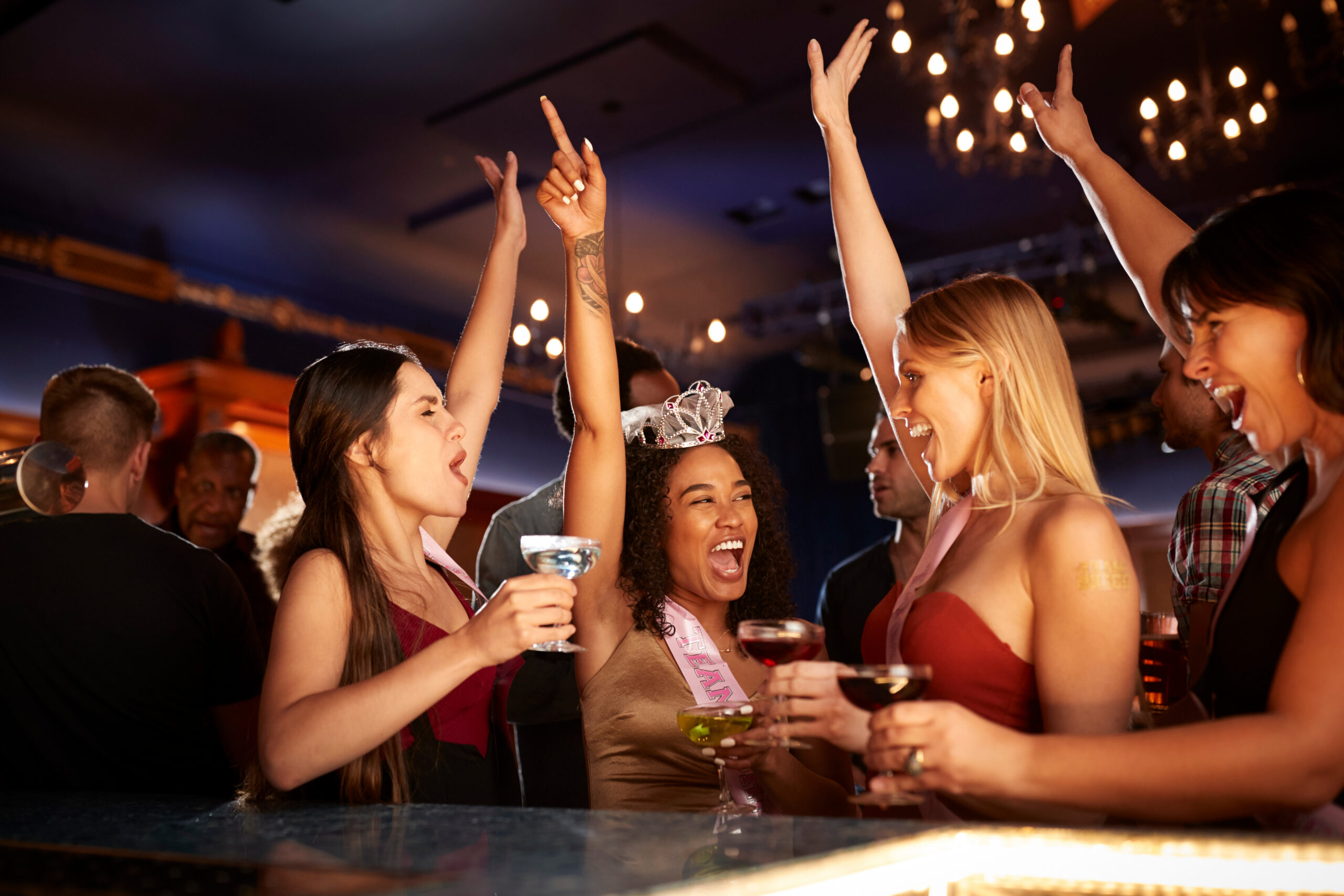 Bride-to-be celebrating her bachelorette party with friends and cocktails at the bar, wearing a tiara and sash